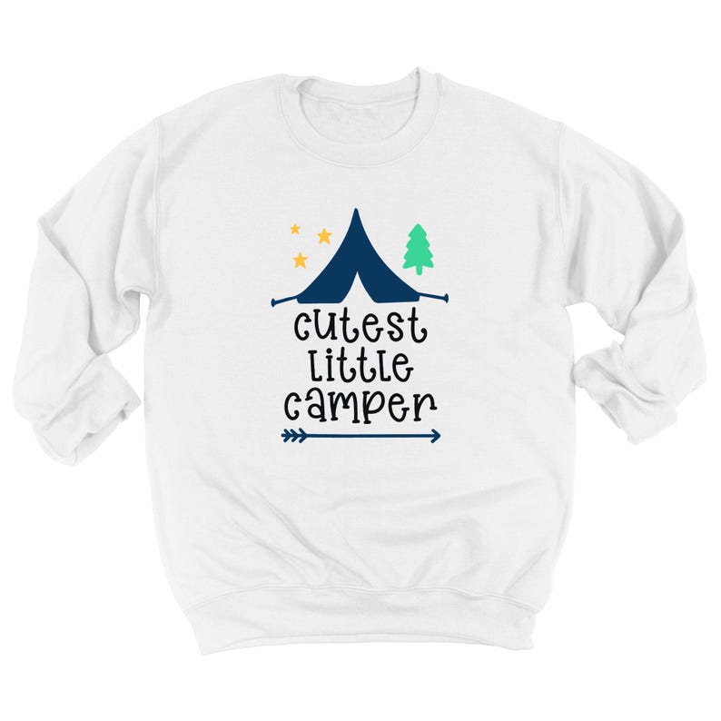 May include: White sweatshirt with a navy blue tent graphic, a green tree, and yellow stars. The text "cutest little camper" is below the tent, with a blue arrow. A cozy, long-sleeved top for camping enthusiasts.
