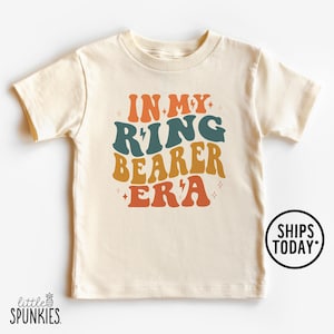 May include: Cream-colored toddler t-shirt with the text "IN MY RING BEARER ERA" in retro orange, teal, and brown lettering. The shirt has a round neck and short sleeves. A small logo is in the bottom left corner, and a circular graphic that says "SHIPS TODAY" is on the right.