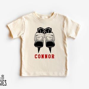 May include: Cream-colored t-shirt featuring a black graphic of two ice skates with white laces. The name "CONNOR" is printed in red below the skates. The shirt has short sleeves and a crew neck.