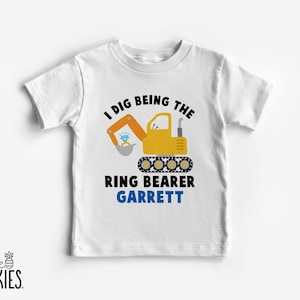May include: White t-shirt with a yellow excavator graphic holding a diamond ring. Text reads "I DIG BEING THE RING BEARER" above the name "GARRETT" in blue. The shirt has a small "little SPUNKIES" logo.