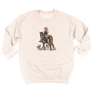 Puede incluir: Sudadera gris claro con una ilustraci&oacute;n de una persona montando a caballo. El jinete lleva un sombrero rosa y vaqueros azules. Un cactus y otras plantas del desierto est&aacute;n en primer plano. La sudadera tiene mangas largas.