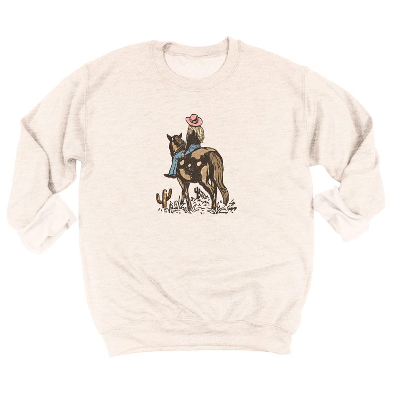 Puede incluir: Sudadera gris claro con una ilustraci&oacute;n de una persona montando a caballo. El jinete lleva un sombrero rosa y vaqueros azules. Un cactus y otras plantas del desierto est&aacute;n en primer plano. La sudadera tiene mangas largas.
