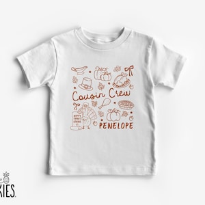 May include: A white short-sleeved t-shirt featuring a brown line-art graphic of Thanksgiving-themed icons including a turkey, pumpkins, a pie, and a gravy boat. The text reads Cousin Crew and PENELOPE, with Happy Thanksgiving on a scroll.