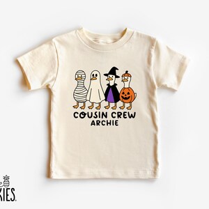May include: A cream-colored short-sleeve t-shirt featuring a graphic of four ducks in Halloween costumes: a mummy, a ghost, a witch, and a pumpkin. Below the ducks, the text reads COUSIN CREW ARCHIE in black, rounded, sans-serif lettering.