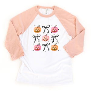 May include: A white raglan shirt with peach-colored three-quarter length sleeves. The front features a repeating pattern of orange and pink jack-o'-lantern pumpkins, black bows, and small black stars printed in a grid-like arrangement.
