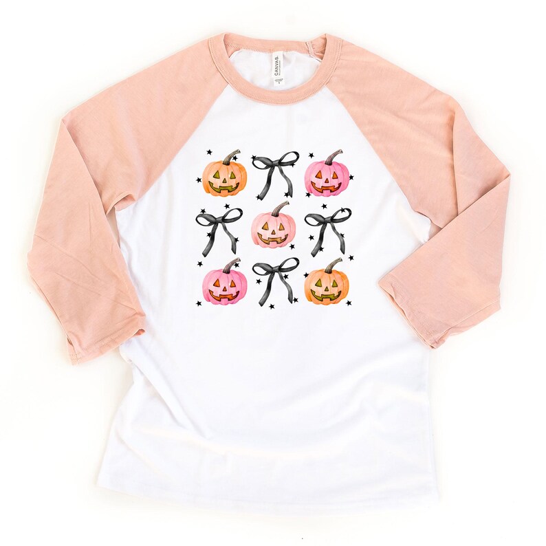 May include: A white raglan shirt with peach-colored three-quarter length sleeves. The front features a repeating pattern of orange and pink jack-o'-lantern pumpkins, black bows, and small black stars printed in a grid-like arrangement.