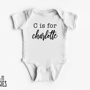 May include: A white short-sleeved baby bodysuit featuring the black printed text C is for charlotte. The text is displayed in a combination of a simple typewriter-style font for the first line and a cursive script font for the name.