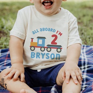 May include: A cream-colored short-sleeve t-shirt featuring a graphic of a colorful train with the text "all aboard I'M 2" in blue and red, and the name "BRYSON" printed in blue below the train illustration on the front of the garment.