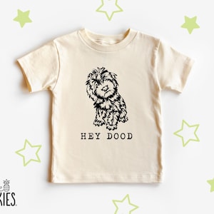 May include: A cream-colored short-sleeve t-shirt featuring a black graphic illustration of a shaggy dog in the center. Below the dog, the text reads HEY DOOD in a bold, sans-serif font. The shirt has a crew neckline and a straight hem.