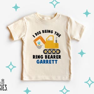 May include: Cream-colored toddler t-shirt with the text "I DIG BEING THE RING BEARER GARRETT". The design features a yellow excavator scooping a diamond ring. The shirt has short sleeves and a crew neck.
