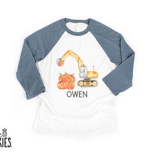 May include: A white raglan t-shirt with blue three-quarter length sleeves. The front features a watercolor illustration of a yellow excavator lifting a pumpkin, with a pile of pumpkins below and the name OWEN printed in black capital letters.