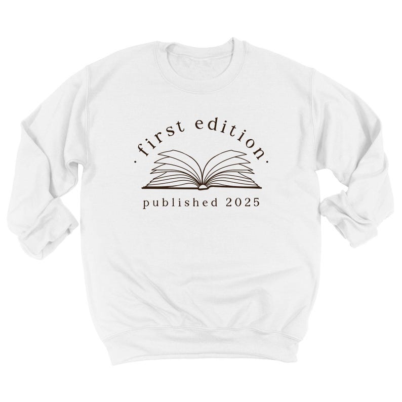 May include: A white long-sleeve sweatshirt featuring a black graphic of an open book centered on the chest. Above the book, the text reads first edition in a curved font, and below the book, the text reads published 2025 in a simple font.