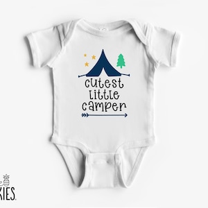 May include: White baby onesie with a navy blue tent, yellow stars, and a green tree graphic. The text "cutest little camper" is printed in black with a blue arrow. The brand logo "little SPUNKIES" is at the bottom.