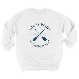 May include: White sweatshirt with a navy blue design featuring crossed oars, waves, and the text "life is better at custom text". The design is centered on the front of the long-sleeved crew neck sweatshirt.