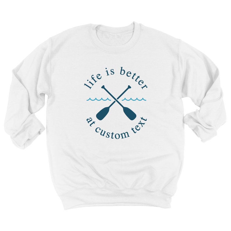 May include: White sweatshirt with a navy blue design featuring crossed oars, waves, and the text "life is better at custom text". The design is centered on the front of the long-sleeved crew neck sweatshirt.