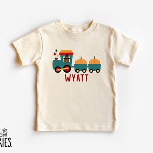 May include: A cream-colored short-sleeve t-shirt featuring a graphic of a teal train engine with a turkey wearing a hat, pulling two cars loaded with orange pumpkins. The name WYATT is printed in red capital letters below the train graphic.