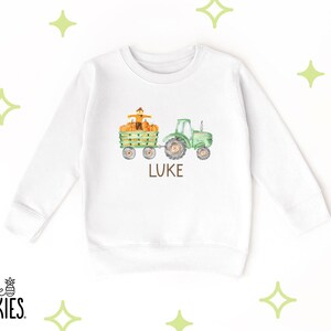 May include: A white long-sleeve sweatshirt featuring a watercolor graphic of a green tractor pulling a wagon filled with orange pumpkins and a scarecrow. The name LUKE is printed in brown capital letters below the tractor illustration.