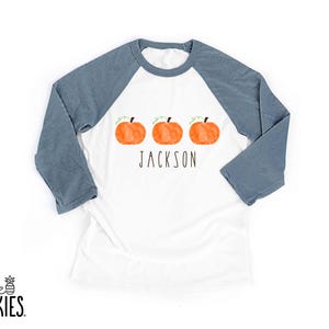 May include: A white raglan shirt with three-quarter length slate blue sleeves. The front features three watercolor-style orange pumpkins in a horizontal row above the name JACKSON printed in a simple, thin, black sans-serif font.
