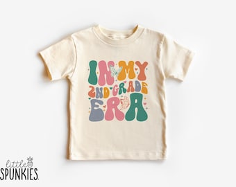 Könnte beinhalten: Cremefarbenes T-Shirt mit dem Text "IN MY 2ND GRADE ERA" in farbenfrohen Buchstaben im Retro-Stil. Das Design enthält Schmetterlings- und Herzgrafiken. Das Markenlogo "little SPUNKIES" befindet sich unten links.