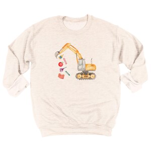 Puede incluir: Sudadera gris claro con una ilustraci&oacute;n en acuarela de una excavadora amarilla con un cuchar&oacute;n negro. La excavadora levanta &uacute;tiles escolares, incluyendo una manzana roja, tijeras y un libro. La sudadera tiene cuello redondo y mangas largas.