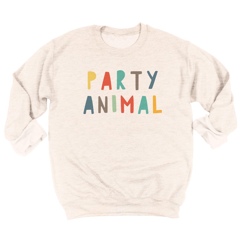 May include: A heathered beige crewneck sweatshirt featuring the text PARTY ANIMAL printed across the chest in a multi-colored, hand-drawn style font. The letters are colored in shades of yellow, red, teal, brown, and blue, set against the fabric.