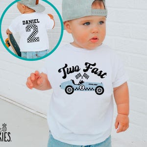 May include: A white t-shirt featuring a graphic of a light blue race car with the text Two Fast and checkered flags. An inset shows the back of the shirt with the name Daniel and the number 2 in a checkered pattern print on the back.