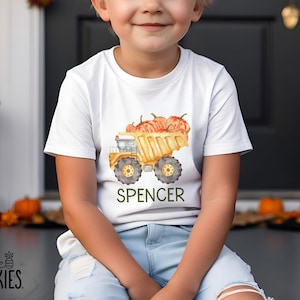 May include: A white short-sleeved t-shirt featuring a watercolor illustration of a yellow dump truck carrying orange pumpkins in its bed. The name SPENCER is printed in dark green capital letters below the truck graphic on the front of the shirt.