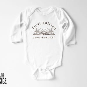 May include: A white long-sleeve baby bodysuit featuring a black graphic of an open book. Above the book, the text reads first edition, and below the book, the text reads published 2027. The garment has a crew neckline and snap closures at the bottom.