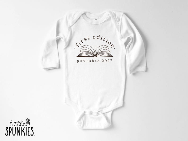 May include: A white long-sleeve baby bodysuit featuring a black graphic of an open book. Above the book, the text reads first edition, and below the book, the text reads published 2027. The garment has a crew neckline and snap closures at the bottom.