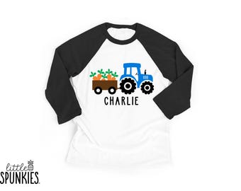 May include: A white baseball-style t-shirt with black three-quarter length sleeves. The front features a graphic of a blue tractor pulling a brown trailer filled with orange carrots and green tops. The name CHARLIE is printed in black below.