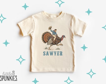 May include: A cream-colored short-sleeve t-shirt featuring a graphic of a person riding a turkey while holding a hat. The text "SAWYER" is printed in blue block letters below the graphic. The design includes brown, blue, and red colors.