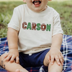 Op de afbeelding: Een cr&egrave;mekleurig T-shirt met de naam "CARSON" in groene en bruine letters met een voetbalthema. Het kind draagt een marineblauwe korte broek en zit op een blauw-rood geruite deken. De foto is buiten genomen.
