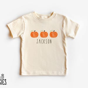 May include: A cream-colored short-sleeve t-shirt featuring a graphic of three orange pumpkins with green stems and vines. Below the pumpkins, the name JACKSON is printed in a thin, black, sans-serif font. The shirt has a crew neckline.