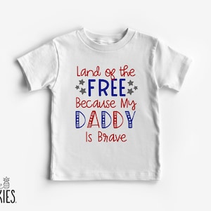 May include: A white short-sleeve t-shirt featuring red, blue, and grey text that reads Land of the FREE Because My DADDY Is Brave, accented with small grey stars and red, white, and blue striped patterns within the letters of the word DADDY.