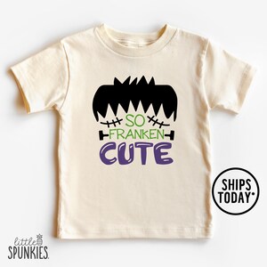 May include: Cream-colored toddler t-shirt with a black Frankenstein's monster hair graphic and the words "SO FRANKEN CUTE" in green and purple. A round sticker with the words "SHIPS TODAY" is on the bottom right.