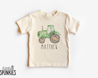 May include: A cream-colored short-sleeve t-shirt featuring a watercolor-style illustration of a green tractor in the center. Below the tractor, the name MATTHEW is printed in black capital letters. The brand logo little SPUNKIES is visible.