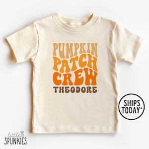May include: Cream-colored short-sleeved t-shirt with the text "PUMPKIN PATCH CREW THEODORE" in orange and brown retro font. A small round sticker on the right says "SHIPS TODAY". The shirt has a crew neck and is made of soft material.