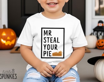 Puede incluir: Una camiseta blanca de manga corta con un borde rectangular negro que contiene el texto negro "MR STEAL YOUR PIE" y un gráfico de un trozo de tarta marrón con nata montada blanca, combinada con unos vaqueros azul claro desgastados.