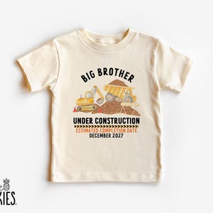 May include: A cream-colored short-sleeve t-shirt featuring a graphic of yellow construction vehicles, a pile of dirt, and the text "BIG BROTHER UNDER CONSTRUCTION ESTIMATED COMPLETION DATE DECEMBER 2027" printed in black and orange lettering.