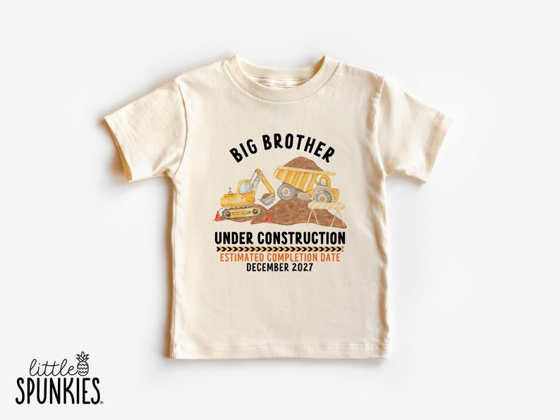 May include: A cream-colored short-sleeve t-shirt featuring a graphic of yellow construction vehicles, a pile of dirt, and the text "BIG BROTHER UNDER CONSTRUCTION ESTIMATED COMPLETION DATE DECEMBER 2027" printed in black and orange lettering.