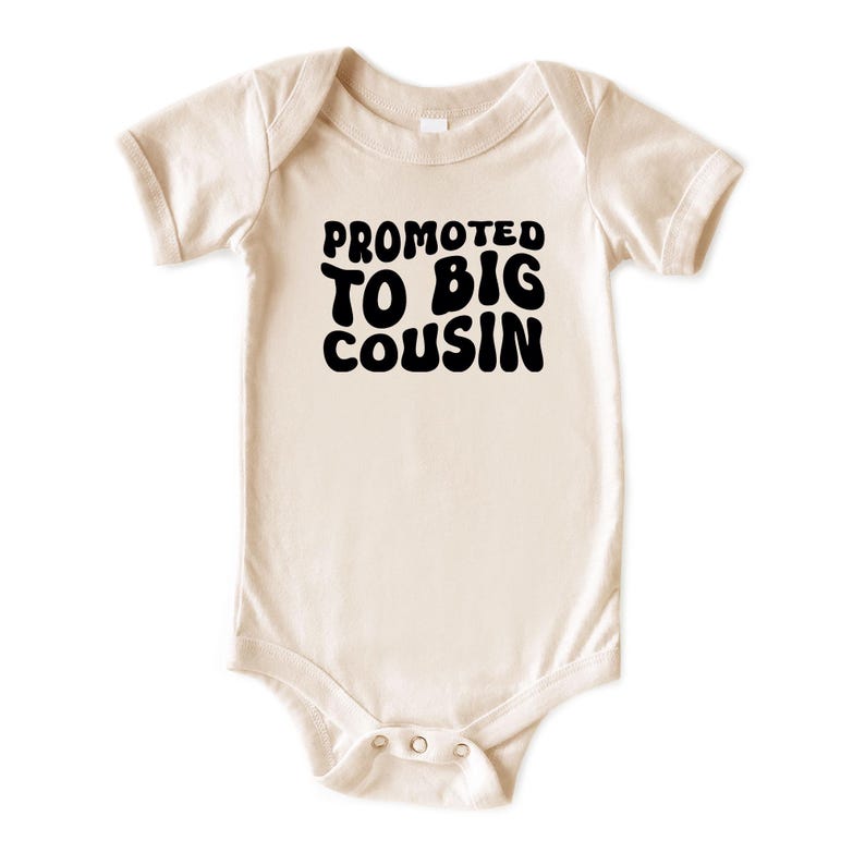 Może przedstawiać: Kremowe body dla niemowląt z kr&oacute;tkimi rękawami. Tekst "PROMOTED TO BIG COUSIN" jest wydrukowany pogrubioną, czarną czcionką bąbelkową z przodu. Body ma zatrzaski na dole.