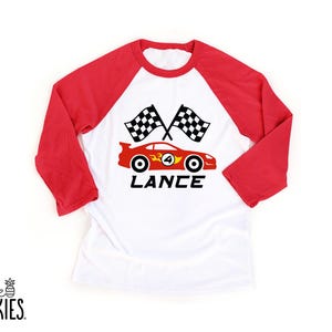 May include: A white baseball-style shirt with red three-quarter length sleeves. The front features a graphic of a red race car with the number 4, two checkered flags, and the name LANCE printed in black text below the car design.