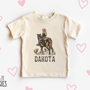 Puede incluir: Camiseta color crema con un gr&aacute;fico de una persona montando a caballo, un cactus y la palabra "DAKOTA". El dise&ntilde;o utiliza tonos marrones, azules y rosas. La camiseta tiene mangas cortas y cuello redondo. El logo de la marca "little SPUNKIES" est&aacute; abajo.