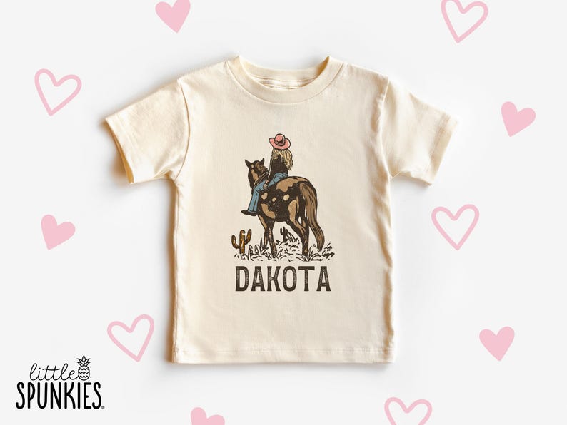 Puede incluir: Camiseta color crema con un gr&aacute;fico de una persona montando a caballo, un cactus y la palabra "DAKOTA". El dise&ntilde;o utiliza tonos marrones, azules y rosas. La camiseta tiene mangas cortas y cuello redondo. El logo de la marca "little SPUNKIES" est&aacute; abajo.