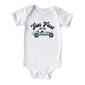 May include: A white short-sleeved bodysuit featuring a graphic of a light blue vintage race car with the number two on the side. Above the car, the text Two Fast is printed in a black distressed font, accompanied by two small checkered flags.