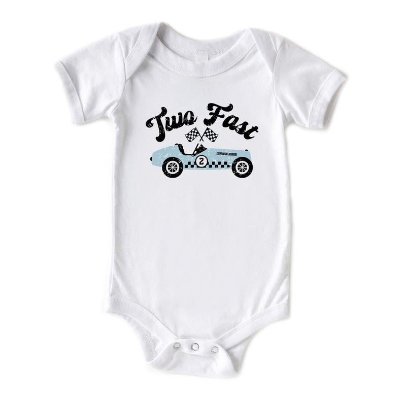 May include: A white short-sleeved bodysuit featuring a graphic of a light blue vintage race car with the number two on the side. Above the car, the text Two Fast is printed in a black distressed font, accompanied by two small checkered flags.