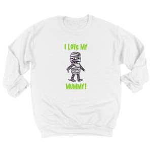 May include: A white long-sleeve sweatshirt featuring a black and white line drawing of a mummy character centered on the chest. Above and below the mummy, the text reads I LOVE MY MUMMY! in a bright lime green, dripping, stylized font.
