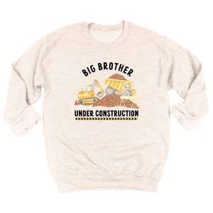 May include: A light beige crewneck sweatshirt featuring a graphic of a yellow excavator and dump truck on a dirt pile. The text reads BIG BROTHER above the vehicles and UNDER CONSTRUCTION below, with a black and yellow hazard stripe pattern.