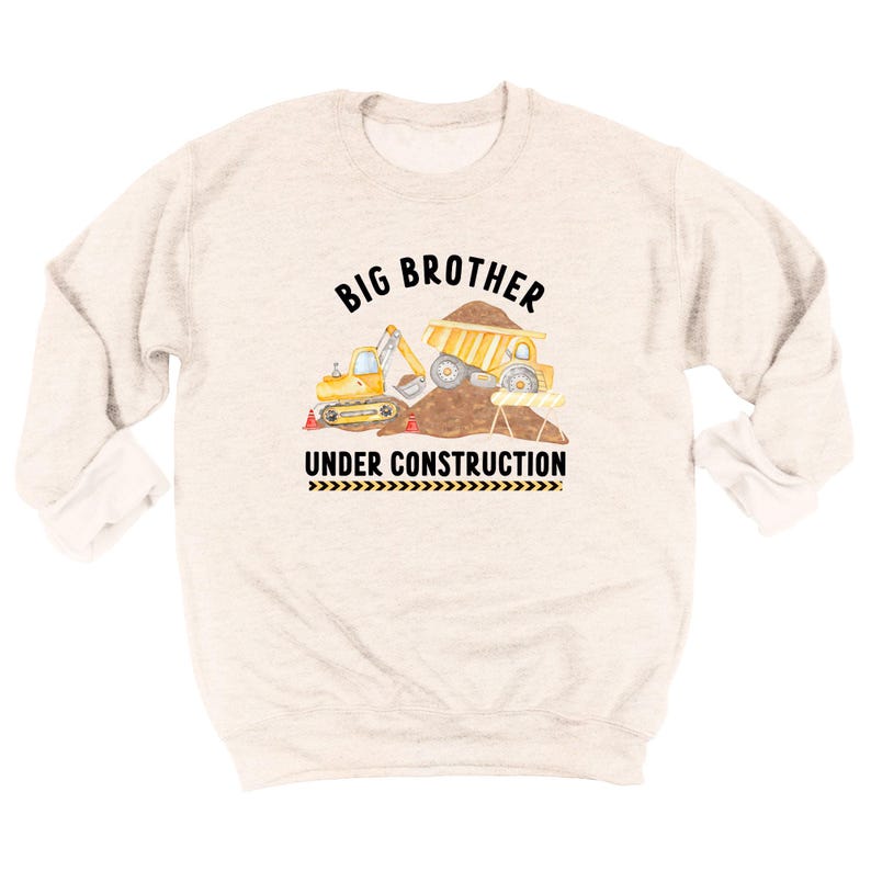 May include: A light beige crewneck sweatshirt featuring a graphic of a yellow excavator and dump truck on a dirt pile. The text reads BIG BROTHER above the vehicles and UNDER CONSTRUCTION below, with a black and yellow hazard stripe pattern.