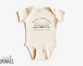 Puede incluir: Body de bebé color crema con mangas cortas. Presenta un gráfico negro de un libro abierto con las palabras "first edition. published 2026" en un diseño circular. El logotipo de la marca "little SPUNKIES" está en la parte inferior.